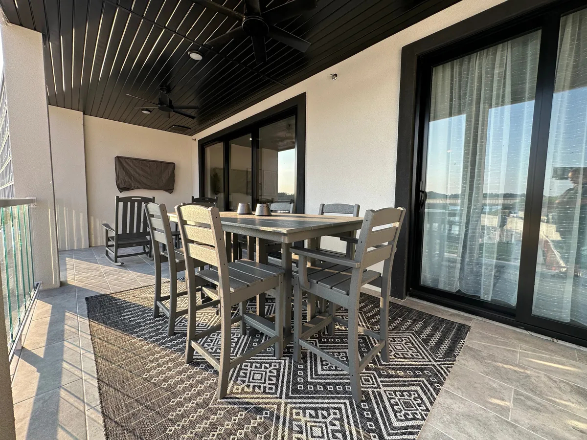 A spacious, covered outdoor dining area featuring a dining table with chairs, a rocking chair, and sliding glass doors leading to the interior. The area rug and relaxed setting overlook the lake, offering a perfect spot for meals or evening gatherings in the fresh air.