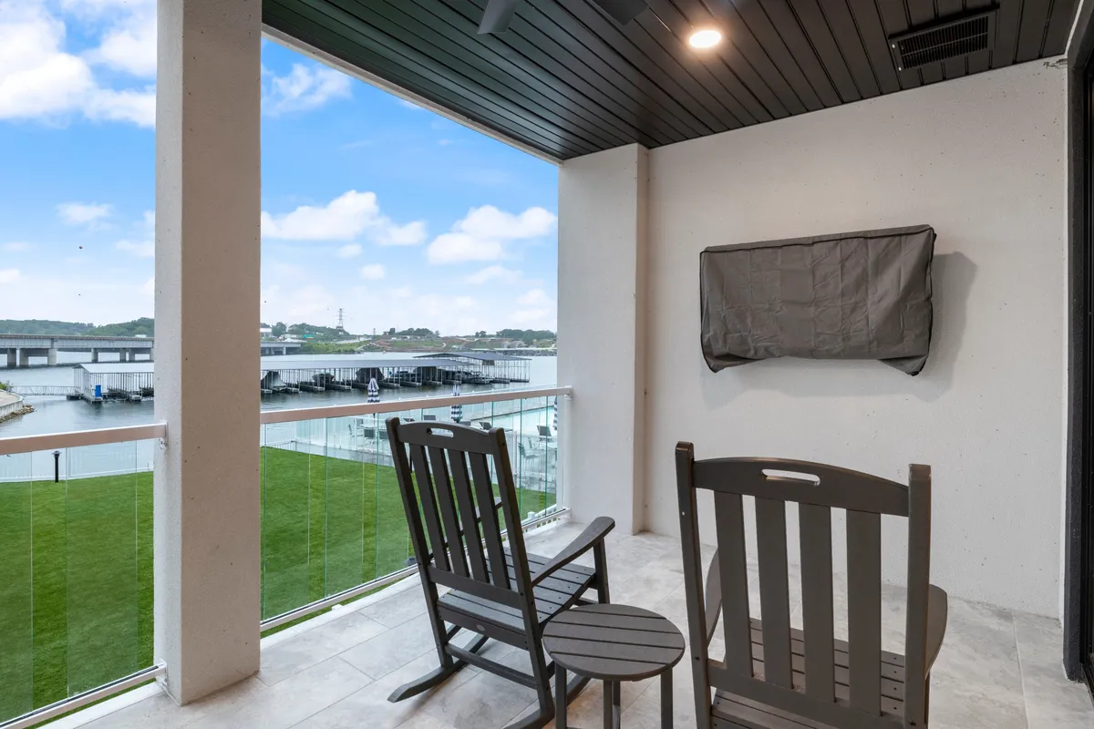 This exterior image captures a serene lakeside setting with rocking chairs facing the shimmering water, a grassy area, and a dock in the distance. The arrangement invites guests to relax, sip coffee, or simply admire the tranquil views.