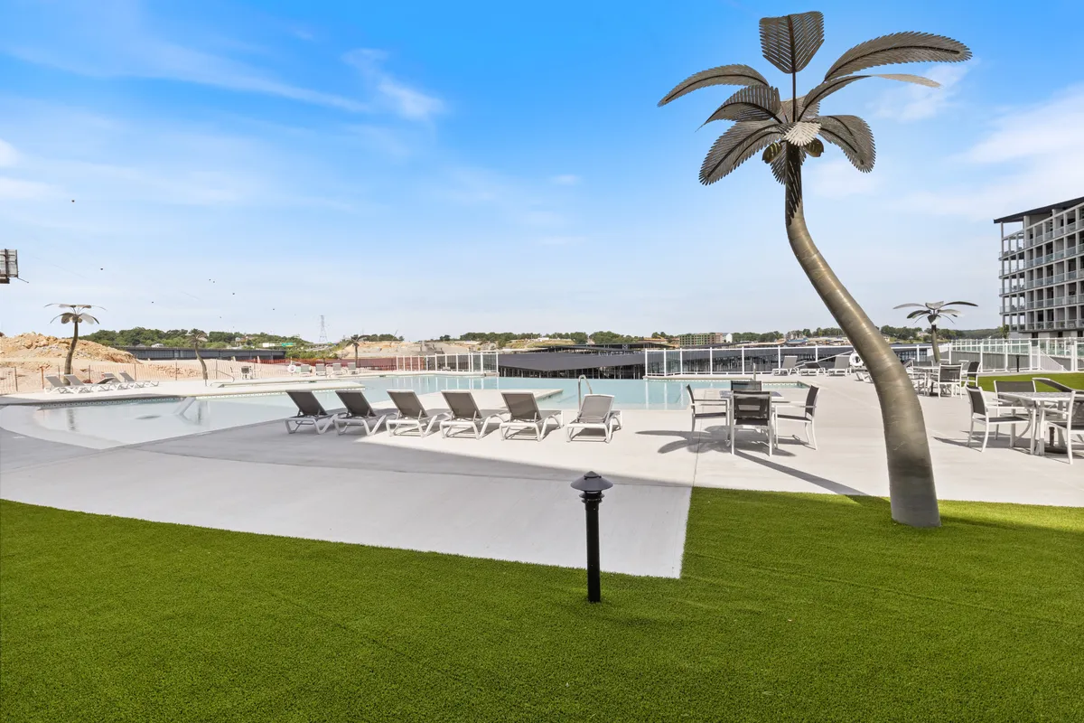 A vibrant swimming pool area surrounded by lounge chairs, a decorative palm tree, and a well-maintained green lawn. The pool is centrally located within the condo property, with plenty of space for families to relax and children to play.