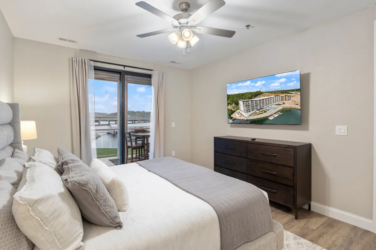A serene bedroom featuring a plush bed with soft pillows and linens, a large window letting in natural light and offering a lovely view, a mounted TV for entertainment, and a dark wooden dresser providing ample storage. The overall ambiance is peaceful and inviting, ideal for relaxation.