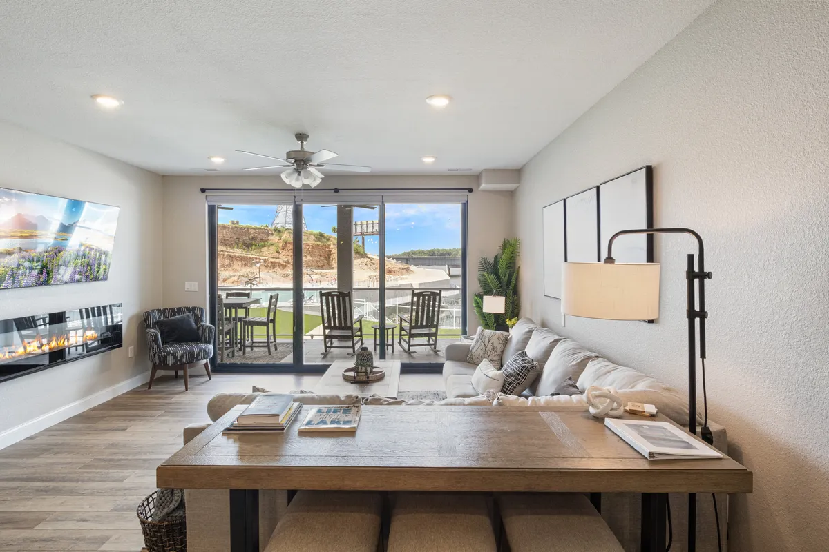 This image displays a spacious open-concept interior with large glass doors that lead to an exterior patio, a modern sofa, a central dining table ideal for group meals, a decorative fireplace, and vibrant wall art. The natural light streaming in through the glass doors accentuates the seamless transition between indoor and outdoor living.
