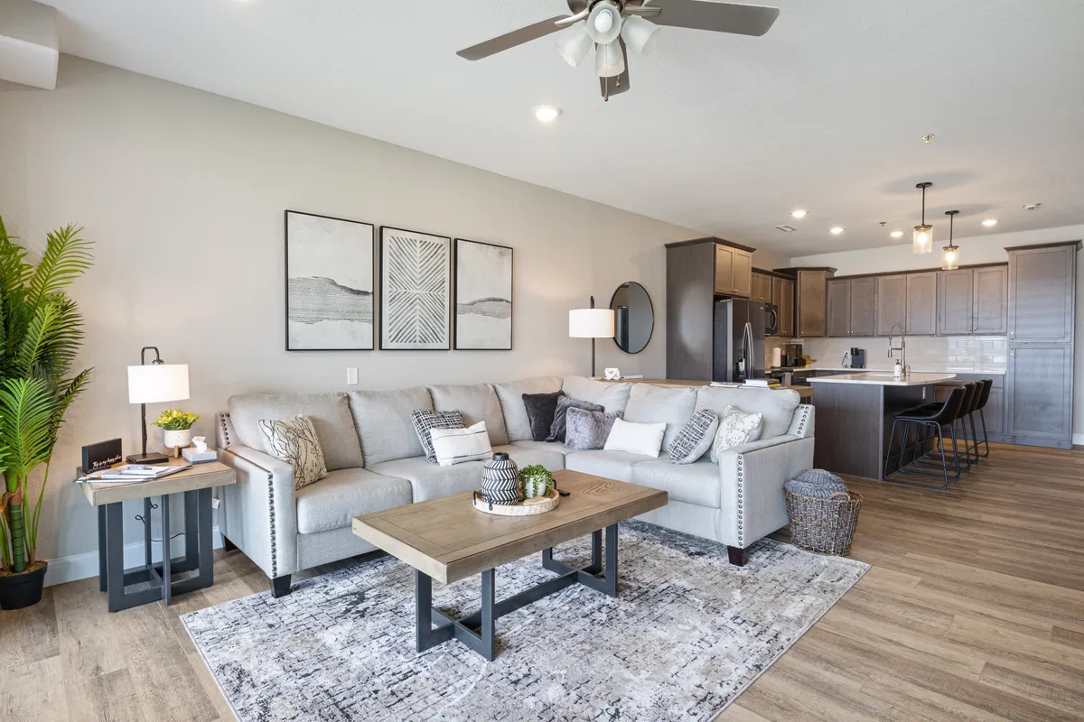 A cozy interior living space featuring a plush sofa, coffee table, stylish artwork, and an open view of the kitchen area. The arrangement suggests comfort and convenience, inviting relaxation or socialization with easy access to household essentials.