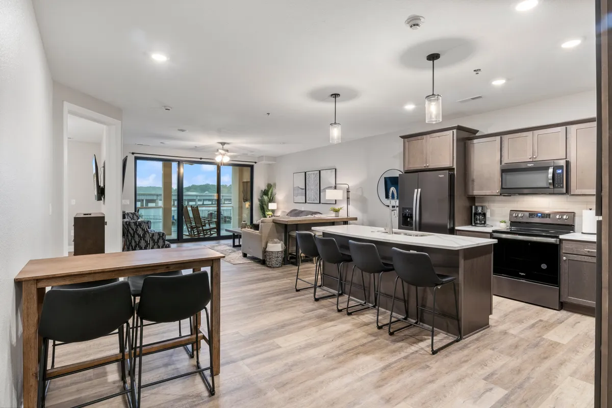 This image features a contemporary kitchen island with bar stools, an adjoining dining area, large windows that flood the room with light, and direct access to a balcony. The arrangement encourages both casual and formal dining, while the open sightlines promote togetherness.